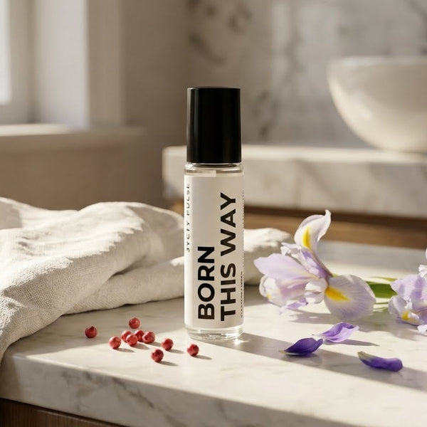 Roller bottle labeled 'Born This Way' on a marble surface with flowers and a towel.