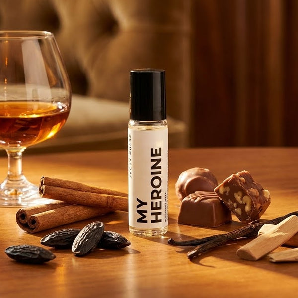 Bottle of 'My Heroine' perfume oil with a glass of amber liquid, cinnamon sticks, and chocolate truffles on a wooden surface. Long lasting and vegan oil